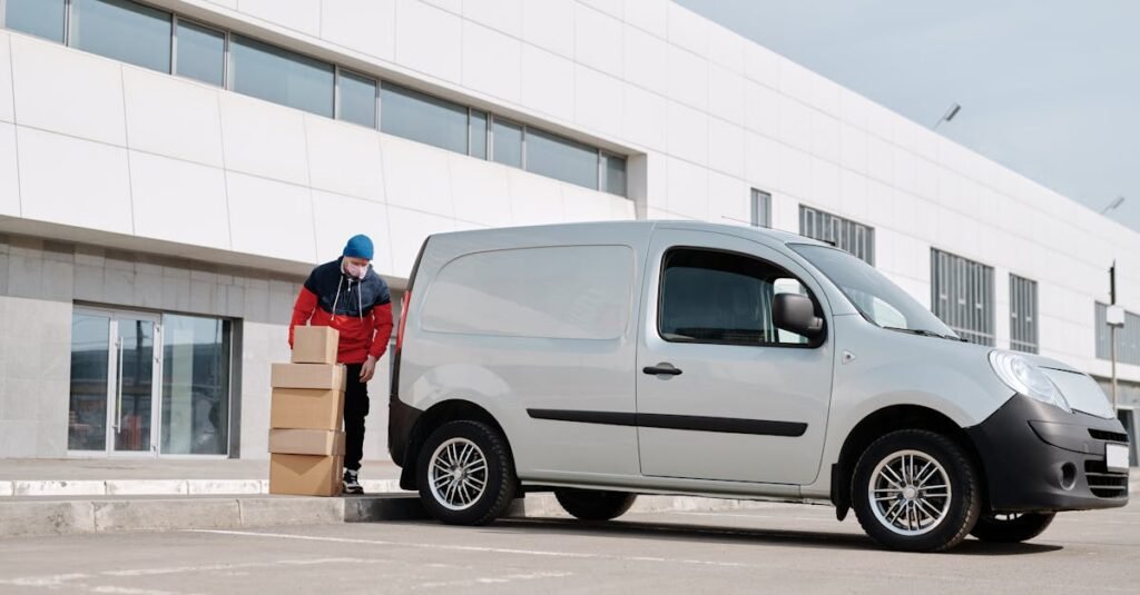 A delivery driver wearing a face mask unloads packages from a van outside a modern building.