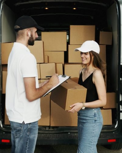 A delivery person hands a package to a woman from a van full of parcels outdoors.